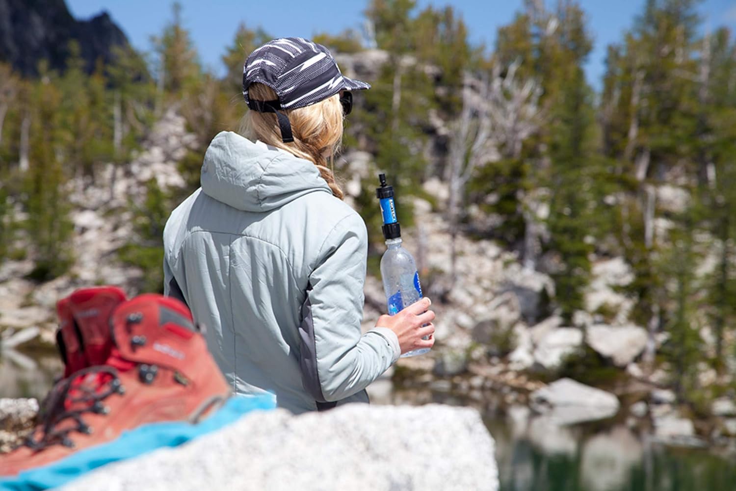 A person using a Portable Water Filter to fill a bottle from a pristine alpine lake, with hiking boots and rugged terrain in the foreground.